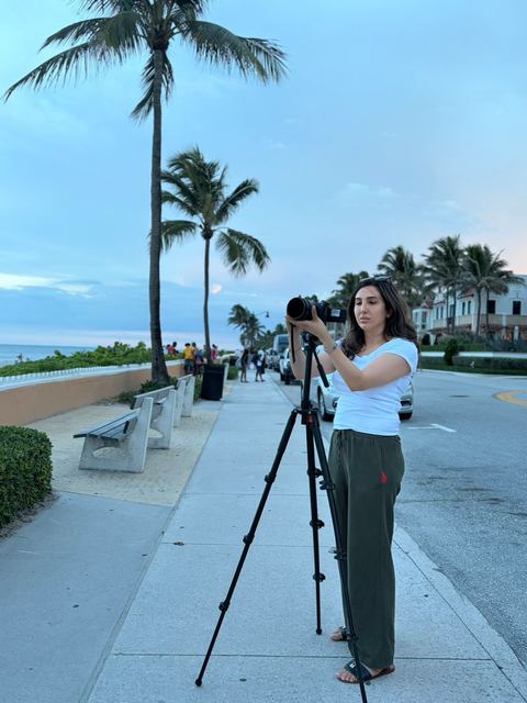 Haya Al Badarneh setting up a camera on a tripod on a palm-lined promenade.